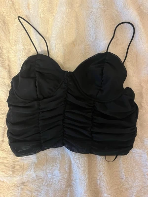 Fest topp svart  - Säljer en svart croppad korsettopp från Gina Tricot i storlek S. Toppen har tunna axelband, rynkade detaljer framtill och mesh-liknande material som ger en cool look. Perfekt att styla med höga jeans eller kjol för en trendig outfit.