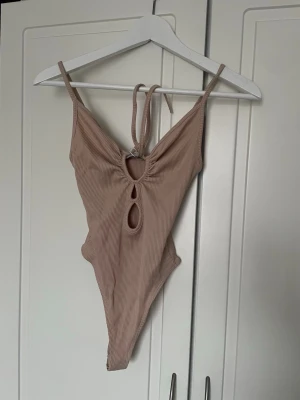 Beige ribbad bodysuit från Bershka - Säljer en helt oanvänd beige ribbad body från Bershka i storlek XS. (Prislapp finns kvar). Bodysuiten har smala axelband, knytning fram och två snygga cut-outs vid bröstet. Materialet är stretchigt och följsamt. Köpt utomlands men var för liten för mig därav att jag nu säljer den