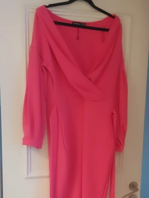 Knallrosa off sholders klänning från Boohoo - Säljer en off sholders, knallrosa klänning från Boohoo med omlott-ringning och långa ärmar. Klänningen har ett bälte i midjan och är tillverkad i ett mjukt, lätt syntetmaterial. Perfekt för dig som vill sticka ut med färg och stil.