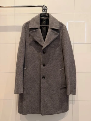 Grå rock från These Glory Days - Snygg grå rock med klassisk krage och tre knappar framtill från These Glory Days. Jackan har diskreta fickor på sidorna och en bröstficka. Yttermaterialet är ull och insidan är fodrad med svart satin för extra komfort. Jag som haft den är 183cm. Nypris 2000kr, mitt pris 500kr