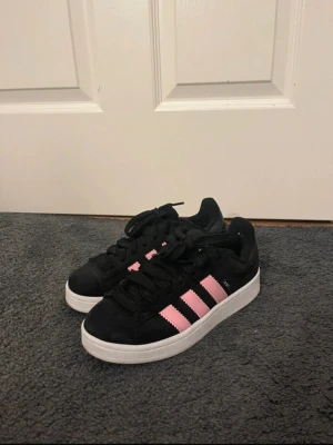 Adidas Campus sneakers svart/rosa - Säljer ett par Adidas Campus sneakers i svart mocka med klassiska rosa ränder på sidorna och vit platt sula. Skorna har rund tå och snörning framtill. Perfekt för dig som gillar stilrena och färgglada sneakers med retrovibe. Köpta för 1000 kroner!