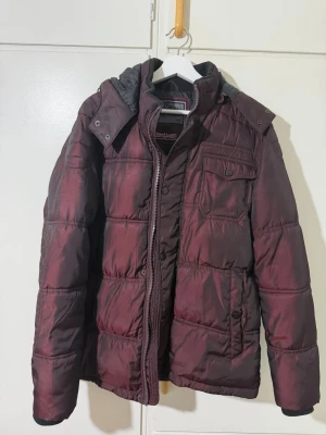 Vinröd pufferjacka från Lee Cooper - Säljer en vinröd pufferjacka från Lee Cooper med huva och flera fickor framtill. Jackan har quiltad design, dragkedja och knappar. Perfekt för kalla dagar och har en snygg, mörk vinröd ton som sticker ut.