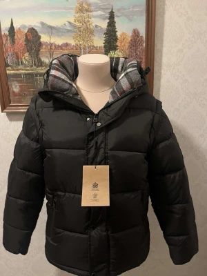 Svart pufferjacka från Burberry - Svart pufferjacka från Burberry med stor huva och rutigt foder. Jackan har en tydlig Burberry London England-patch på ärmen, dragkedja framtill och justerbar huva med spänne. Perfekt för kalla dagar och har en schysst, modern look. Pris kan diskuteras 