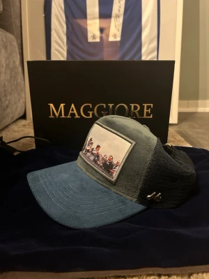 Blå och grå keps från Maggiore - Säljer en snygg keps från Maggiore i blått och grått med mesh på sidorna och baksidan. Framsidan har en patch med ett foto av ett gäng personer. Skärmen är i blå mocka och kepsen har justerbart spänne bak. Perfekt accessoar för en cool streetstil.