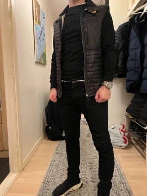 Svart quiltad dunväst från Parajumpers - Säljer en svart quiltad dunväst från Parajumpers med dragkedja framtill och bröstficka med zip. Västen har en snygg ståkrage och ett litet gult spänne vid halsen. Perfekt att slänga över en hoodie eller tröja för extra värme.