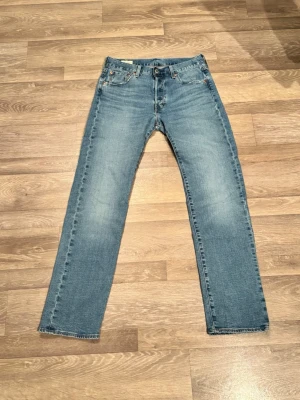 Levi's 501 ljusblå jeans W29 L32 - Klassiska Levi's 501 jeans i ljusblå tvätt med raka ben och femficksmodell. Jeansen har knappgylf, ikonisk läderpatch bak och röd Levi's-tag på bakfickan. Tillverkade i slitstarkt bomullsjeansmaterial. Perfekta för en avslappnad och tidlös look.