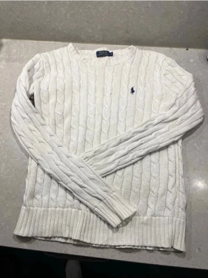 Vit kabelstickad tröja Polo Ralph Lauren - Säljer en vit kabelstickad tröja från Polo Ralph Lauren i storlek XS. Tröjan har långärmad design, rund hals och klassisk broderad logga på bröstet. Perfekt för dig som gillar stilrena och tidlösa plagg med snygg struktur.