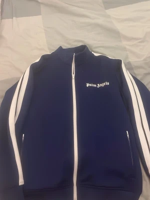 Blå Palm Angels zipper L/M - Säljer en blå track zipper från Palm Angels i storlek L/MJackan har vita ränder längs ärmarna, hel dragkedja framtill och Palm Angels-logga på bröstet. Tillverkad i 100% polyester med ribbade muddar och sportig look.