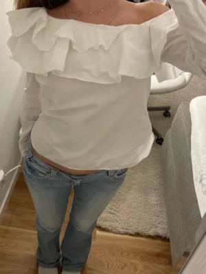 Vit sne shoulder blus med volang - Säljer en vit blus med offshoulder-design och stora volanger längs halsringningen. Blusen har lång ärm och är tillverkad i ett lätt, vävt material som ger en luftig känsla. Perfekt för dig som gillar romantiska detaljer och vill sticka ut lite extra.