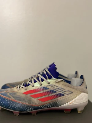 Adidas fotbollsskor blå/röd - Säljer ett par Adidas fotbollsskor med cool design i blått, rött och beige. Skorna har snörning, låg profil och mönstrad ovandel med tydliga Adidas-ränder. Sulan är vit med röda och beige dobbar, perfekt för gräsplan. Ovandelen är i syntetmaterial och har en blå plös. Tvättar innan frakt 