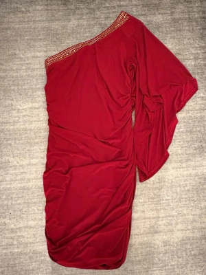Röd oneshoulder klänning med paljetter - Snygg röd oneshoulder klänning med glittrande paljettdetaljer längs halsringningen. Klänningen har en asymmetrisk design med en vid ärm och draperade detaljer.