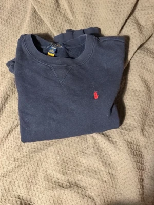 Mörkblå sweatshirt från Polo Ralph Lauren - Klassisk mörkblå sweatshirt från Polo Ralph Lauren med röd broderad logga på bröstet. Tröjan har rund halsringning, ribbade muddar och är tillverkad i mjuk bomull. Perfekt för en avslappnad och stilren look. OBS barnstorleken L