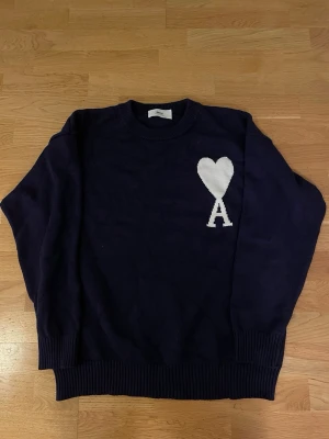 Mörkblå stickad tröja från Ami - Snygg mörkblå stickad tröja från Ami Alexandre Mattiussi med vit hjärt- och A-logga på bröstet. Tröjan har rund hals, ribbade muddar och lång ärm. Perfekt för dig som gillar stilrena detaljer och vill ha något som sticker ut lite.