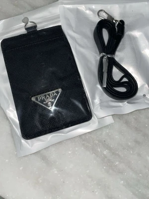 Svart plånbok - Säljer en stilren svart plånbok från P med klassisk trekantig metallogo framtill. Väskan är tillverkad i slitstarkt syntetmaterial och kommer med en matchande svart axelrem med metallspänne. Perfekt för att bära det viktigaste på ett snyggt sätt.