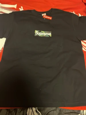 Svart Supreme t-shirt med camo-logga - Supreme t-shirt i svart med klassisk box logo framtill i camouflage-mönster och vit Supreme-text. Rund hals och korta ärmar. Materialet är mjuk bomull, perfekt för streetwear och chill stil. Tröjan är helt ny och jag säljer den för att den inte passade mig 