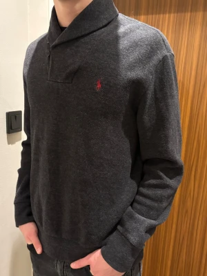 Vintage Polo Ralph Lauren  - Mörkgrå tjocktröja från Polo Ralph Lauren. En rare vintage tröja som har en unik krage med knappar och är gjord i mjuk bomull.                                    Riktigt najs mocka lappar på armbågarna. Tröjan är i väldigt bra skick!                                            Storlek L men sitter som M. Vanligtvis bär jag aldrig L men denna sitter som sagt som en M!