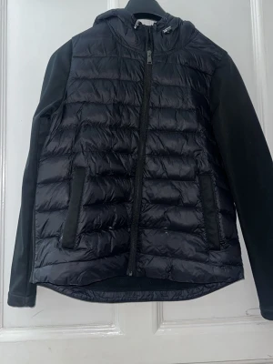 Svart pufferjacka från Polo Ralph Lauren - Svart pufferjacka från Polo Ralph Lauren med huva och dragkedja framtill. Jackan har quiltad framsida, släta ärmar och två sidofickor. Materialet är polyester och jackan har en clean, sportig vibe som funkar till många olika stilar.