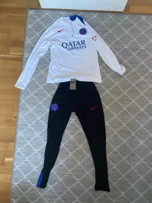 PSG Tracksuit 25/26 Player Version Helt Ny!!! - Säljer ett helt nytt träningsset från Paris Saint-Germain, säsongen 2025/2026. Setet består av en långärmad vit träningströja och svarta träningsbyxor – båda med PSG:s officiella klubbmärke och Nike-logga i rött. Tröjan har quarter-zip och tryckt sponsor “Qatar Airways” på bröstet. Byxorna har elastisk midja med snörning, blå sidodetalj och matchande emblem. 