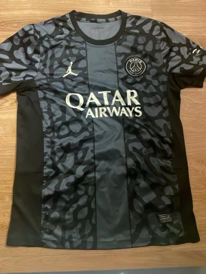 Svart PSG x Jordan matchtröja - Säljer en svart PSG x Jordan matchtröja med grått leopardmönster och vita loggor. Tröjan har Qatar Airways-tryck på bröstet och är tillverkad i ett lätt, syntetiskt material som andas. Perfekt för dig som vill sticka ut på planen eller på stan.