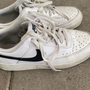 Nike Court Vision vita sneakers - Nike Court Vision sneakers i vitt med svart swoosh på sidan och Nike-logga på plösen och hälen. Skorna har snörning, perforerad tå och platt sula. Tillverkade i syntetmaterial och skinn, klassisk och stilren design som passar till jeans eller shorts.