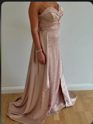 Champagnefärgad långklänning med pärlor - Säljer en elegant långklänning i champagnefärg med smala axelband och v-ringad överdel. Klänningen har pärlbroderier och paljetter på livet samt ett glittrigt mönster på kjolen. Materialet är satin och tyget faller snyggt med en slits framtill. Släppet är avtagbart.
