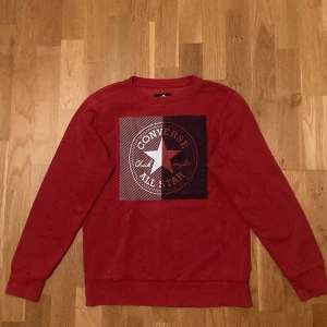 Röd Converse sweatshirt med tryck - Säljer en röd sweatshirt från Converse. Tröjan har ett stort grafiskt tryck med Converse All Star-loggan på bröstet och är långärmad med rund hals. Materialet är mjukt och skönt, perfekt för chill dagar. Passformen är normal och tröjan är tillverkad i bomull. Den är nässtan aldrig använd.