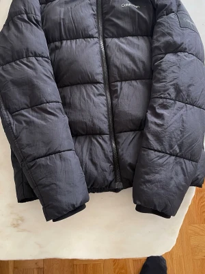 Svart pufferjacka från Calvin Klein - Svart pufferjacka från Calvin Klein i storlek M. Jackan har en stilren design med quiltade sektioner och dragkedja framtill. Perfekt för kalla dagar och har långärmade ärmar. Materialet är syntet och polyester för extra värme.