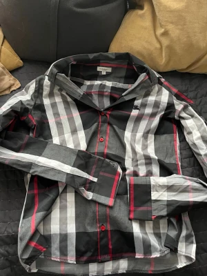 Burburry rutig skjorta - Burberry-inspirerad skjorta – stl XS/S) 🔲🔳 Fick den av min kusin förra året, så jag kan inte garantera att den är äkta – därför säljs den billigt. Passar jättebra i storlek och har riktigt snygga färger med röd detalj och klassiskt rutmönster. Skön kvalitet och stilren look – perfekt till både vardag och fest!