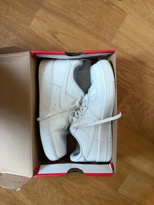 Nike Air Force 1 vita sneakers - Nike Air Force 1 sneakers i helvitt skinn med klassisk rund tå och platt sula. Skorna har snörning och perforerade detaljer på tån för extra ventilation. Kommer i original röd Nike-låda. Perfekt för dig som gillar stilrena och ikoniska sneakers.