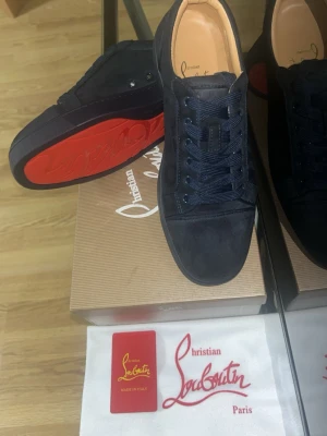 Mörkblå sneakers från Christian Louboutin - Säljer ett par mörkblå sneakers från Christian Louboutin med klassisk röd sula och rund tå. Skorna har snörning och är tillverkade i mocka med diskreta nitar på sidan. Snygg och stilren design med platt sula och exklusiv känsla.