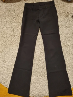 Svarta bootcut kostymbyxor - Snygga svarta kostymbyxor med bootcut och medium midja som man kan vika ner. Byxorna har två bakfickor med lock och knapp, samt knäppning fram med två knappar och dragkedja. Perfekt för dig som gillar klassisk stil med lite extra edge och som man kan ha på finare tillfällen.