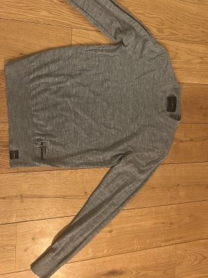 Grå stickad tröja John Richmond - Snygg grå stickad tröja från John Richmond med rund hals och diskret logga vid nederkanten. Tröjan har långärmad design och är tillverkad i mjukt material som passar perfekt för kyliga dagar. Enkel och stilren look som funkar till många outfits.