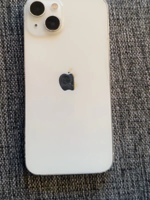 iPhone 13 - Apple iPhone 13 i vitt utförande. Mobilen har dubbla kameror och fungerar som den ska. Skalet visar tydligt slitage och repor, särskilt på sidorna och kring Apple-loggan. Perfekt för dig som vill ha en prisvärd iPhone med full funktionalitet trots kosmetiska defekter.