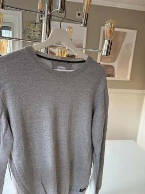 Grå stickad tröja från Seven Seas - Snygg grå stickad tröja från Seven Seas Copenhagen i storlek medium. Tröjan har rund hals, ribbade muddar och är långärmad. Perfekt för dig som gillar enkel och stilren design med mjukt material.