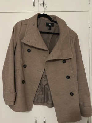 Beige skepparkavaj från H&M strl 40 - Säljer en beige skepparkavaj från H&M i storlek 40. Jackan har dubbelknäppning med svarta knappar, bred krage och är helfodrad. Materialet är mjukt och har en stilren look som passar perfekt till höst och vinter. Snygg att styla med jeans eller boots.