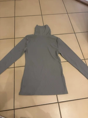 Grå polotopp med lång ärm - Säljer en stilren grå polotopp med lång ärm. Toppen har en hög krage och är ribbad i materialet vilket ger en snygg struktur. Perfekt att styla med jeans eller kjol för en enkel men trendig look.