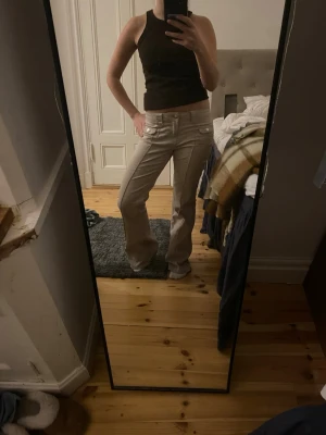 Beige bootcut byxor med fickdetaljer - Säljer ett par snygga beige bootcut byxor med framfickor som har lock och knappar. Byxorna har en lågmidjad passform och markerad söm längs benen. Materialet känns som bomull och de är perfekta för dig som gillar en chill och trendig look.