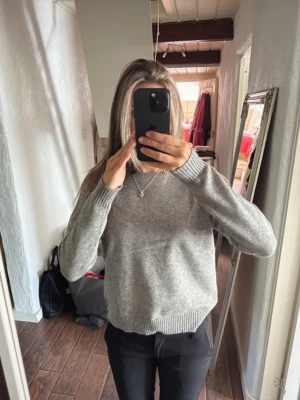 Grå stickad tröja med rund hals - Säljer en enkel och stilren grå stickad tröja med rund hals och ribbade muddar vid ärmslut och nederkant. Tröjan har lång ärm och en normal passform, perfekt att matcha med jeans eller kostymbyxor.