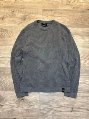 Grå stickad tröja från Pull&Bear - Säljer en grå stickad tröja från Pull&Bear i storlek S. Tröjan har en rund halsringning och ett diskret ribbat mönster över hela plagget. Perfekt för dig som gillar enkel och stilren design med lite struktur. Långa ärmar och mjukt material.