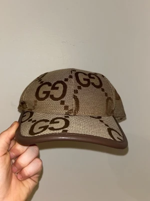  beige-brun keps med GG-mönster - Snygg keps i beige och brunt med det klassiska GG-mönstret över hela kepsen. Kepsen har brunt läder på kanten och justerbar rem bak. Materialet är bomull och polyester, med inslag av elastan. Perfekt för dig som gillar lyxiga accessoarer.