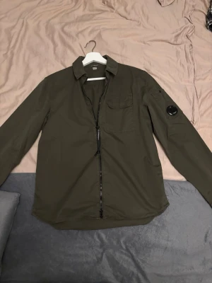Grön overshirt från C.P. Company - Säljer en snygg militärgrön overshirt från C.P. Company med klassisk linsdetalj på ärmen och bröstficka. Jackan har dragkedja framtill. Overshirten är köpt på miinto för ca 5 månader sedan. Säljer den pga att den är lite liten och kommer aldrig till andvändning. Den är i nyskick då den är andvänd (en gång!!)  köptes för 3500kr och nu är den på rea men bara i 2xl. 