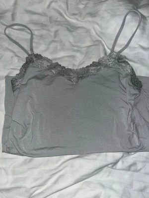 Grå linnetopp med spets från Intimissimi - Säljer en stilren grå linnetopp från Intimissimi i storlek L. Toppen har tunna axelband och v-ringad spetskant upptill som ger en snygg detalj. Materialet är mjukt och följsamt, perfekt för sommaren eller som layering-piece.