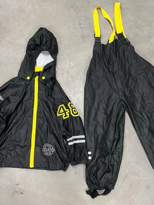 Svart regnjacka med gula detaljer Wear - Svart regnjacka med huva och gula detaljer från Wear by Lindex, storlek 92. Jackan har gula knappar, dragkedja och tryckta siffror på ärmen. Reflexer på både jacka och byxa för extra synlighet. Materialet är syntet och passar perfekt för blöta dagar.