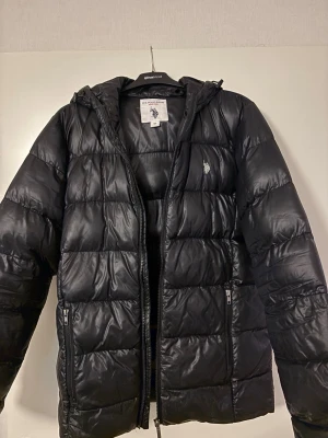 Svart pufferjacka från U.S. Polo Assn. - Svart pufferjacka från U.S. Polo Assn. med huva och broderad logga på bröstet. Jackan har dragkedja framtill och två sidofickor med dragkedja. Materialet är glansigt och känns syntetiskt, perfekt för kalla dagar. Storlek M