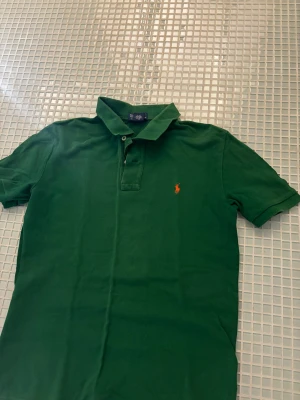 Grön pikétröja från Ralph Lauren - Säljer en klassisk grön pikétröja från Ralph Lauren med orange broderad logga på bröstet. Stilig piké med en unik och ögonfångande färg så man sticker ut. Legat i garderob länge och därmed lite skrynklig men går att lösa enkelt bara att stryka en gång. Storlek S/M