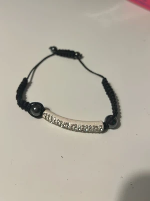 Ett supersnyggt armband  - Bilden visar ett armband med svart snöre och en dekorerad silverfärgad böjd detalj med silver stenar.