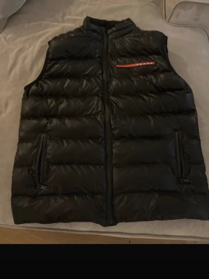 Svart dunväst från Prada - Snygg svart dunväst från Prada med quiltad design och diskret logga på bröstet. Västen har två fickor med dragkedja och hög krage. Materialet är nylon på utsidan och fodrad med dun och vitt ejderdun, vilket gör den både lätt och varm.