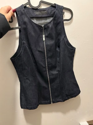 Svart denim korsett-topp från Zara - Säljer en svart korsett-topp från Zara i denim med snygg dragkedja framtill. Toppen är ärmlös och har en figurnära passform som ger en cool siluett. Perfekt att styla med jeans eller kjol för en trendig look.