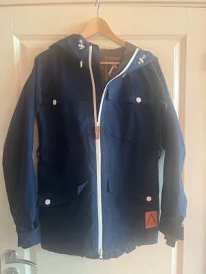 Blå vindjacka från WearColour XS - Blå vindjacka från WearColour i storlek XS med vit dragkedja, huva med snörning och flera praktiska fickor med knappar, funkar till alla väder. Använd ett fåtal gånger. Perfekt för dig som gillar att vara ute och röra på dig.