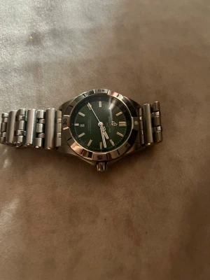 Klocka - Bilderna visar en klocka från Breitling med grön urtavla och metallarmband, men ingen väst finns med på bilderna.